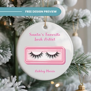 以下が含まれることがあります： ピンクのまつげのデザインと「Santa's Favorite Lash Artist」と「Ashley Marie」の文字が入った、白くて丸いクリスマスオーナメント。オーナメントは、雪の降るクリスマスツリーを背景に、金の紐で吊るされています。