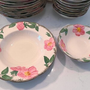 May include: Cream-colored ceramic bowls with pink rose and green leaf designs. The bowls have a scalloped edge and a gray trim. Stacks of matching plates are visible in the background.