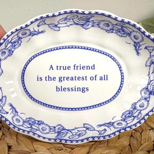 May include: White oval dish with a blue floral border and the text "A true friend is the greatest of all blessings." The dish has a scalloped edge and is likely made of ceramic or porcelain.