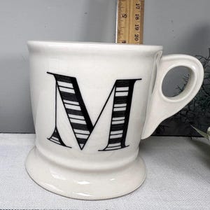 Op de afbeelding: Witte keramische mok met een grote zwarte letter "M" op de voorkant. De letter is gestileerd met horizontale strepen. De mok heeft een afgeronde basis en een gebogen handvat.