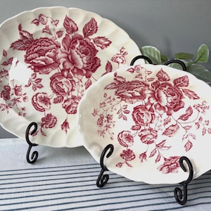 May include: Two cream-colored ceramic plates with a red floral pattern, displayed on black metal stands. The larger plate has a scalloped edge with cut-out details. The floral design features roses and leaves.