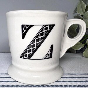 Op de afbeelding: Witte keramische mok met een grote zwarte letter "Z" met een ruitpatroon. De mok heeft een afgeronde basis en een handvat met een uniek, open ontwerp. De mok staat op een gestreept oppervlak.