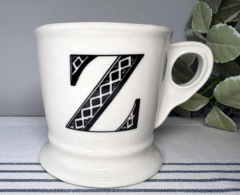 Op de afbeelding: Witte keramische mok met een grote zwarte letter "Z" met een ruitpatroon. De mok heeft een afgeronde basis en een handvat met een uniek, open ontwerp. De mok staat op een gestreept oppervlak.