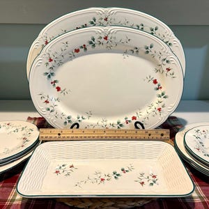 May include: A collection of white ceramic serving dishes with a green trim and a floral design of green leaves and red berries. Includes oval platters, rectangular basketweave tray, and round plates.