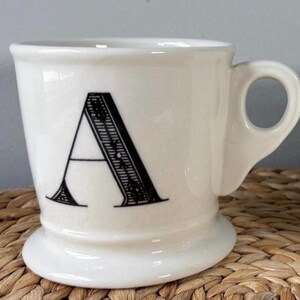 Pu&ograve; includere: Tazza in ceramica color crema con una grande lettera "A" nera sul davanti. La tazza ha una base arrotondata, un manico curvo e un design classico.