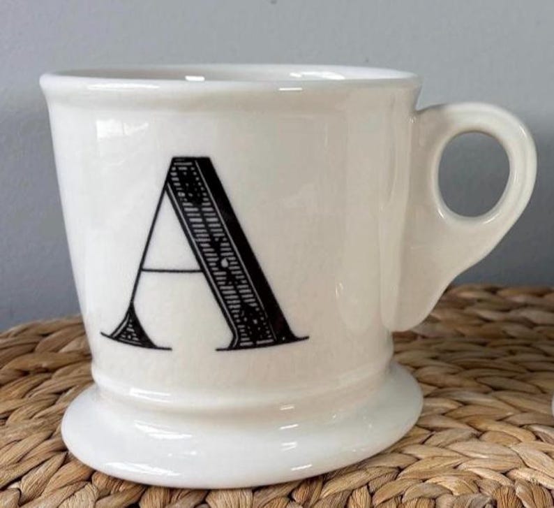 Pu&ograve; includere: Tazza in ceramica color crema con una grande lettera "A" nera sul davanti. La tazza ha una base arrotondata, un manico curvo e un design classico.