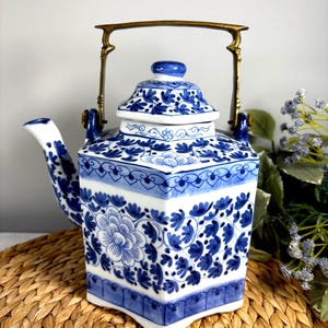 May include: A hexagonal ceramic teapot featuring a white base with intricate blue floral patterns. The teapot includes a matching lid with a blue knob and a metallic brass-colored swing handle attached to two blue ceramic loops on the top rim.