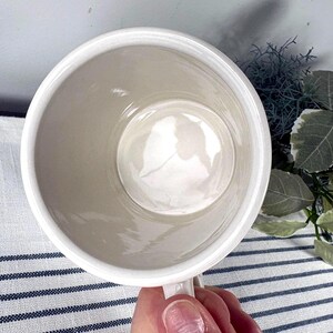 Op de afbeelding: Een witte keramische mok met een handvat, van bovenaf gezien. De binnenkant is lichtbeige van kleur en de rand is wit. De mok wordt door een hand vastgehouden en de achtergrond omvat een gestreepte stof en groen.