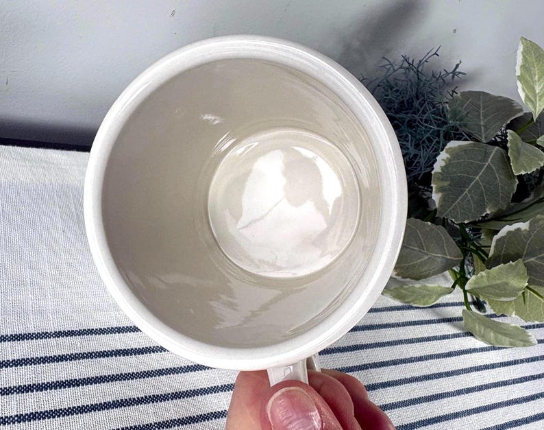 Op de afbeelding: Een witte keramische mok met een handvat, van bovenaf gezien. De binnenkant is lichtbeige van kleur en de rand is wit. De mok wordt door een hand vastgehouden en de achtergrond omvat een gestreepte stof en groen.
