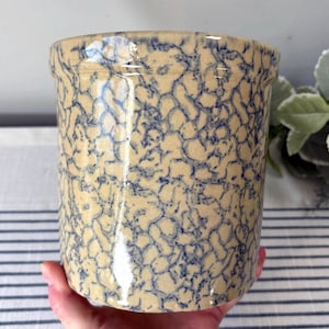 May include: A beige stoneware crock with a blue, web-like pattern. The cylindrical container has a slightly flared rim and is likely used for kitchen storage or decoration. The handmade pottery has a rustic, vintage aesthetic.