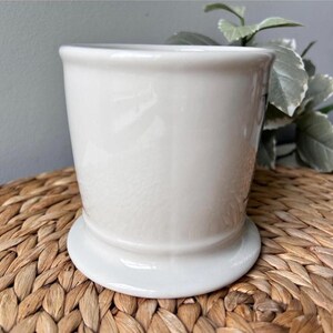 Pu&ograve; includere: Un vaso di ceramica bianco con un corpo arrotondato e un'ampia base circolare. Il vaso ha una finitura liscia e lucida ed &egrave; posto su uno sfondo intrecciato marrone. Il design &egrave; semplice ed elegante, adatto a varie piante.