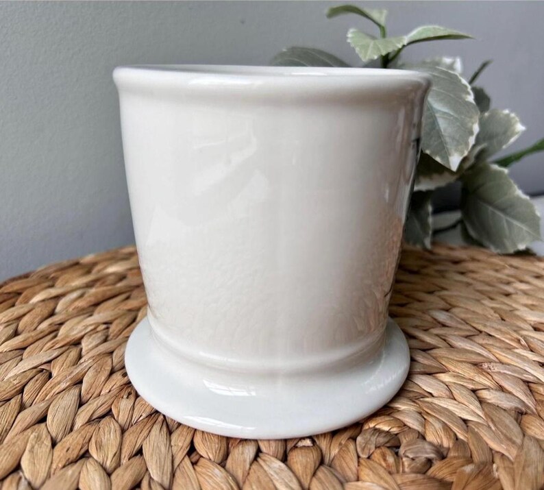 Pu&ograve; includere: Un vaso di ceramica bianco con un corpo arrotondato e un'ampia base circolare. Il vaso ha una finitura liscia e lucida ed &egrave; posto su uno sfondo intrecciato marrone. Il design &egrave; semplice ed elegante, adatto a varie piante.