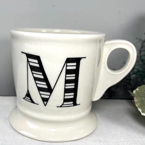 Op de afbeelding: Een witte keramische mok met de zwarte letter "M" met horizontale strepen. De mok heeft een oor en een voet. De achtergrond is neutraal grijs. Geschikt voor koffie of thee.