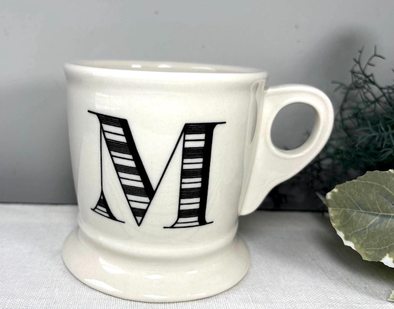 Op de afbeelding: Een witte keramische mok met de zwarte letter "M" met horizontale strepen. De mok heeft een oor en een voet. De achtergrond is neutraal grijs. Geschikt voor koffie of thee.
