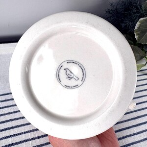Op de afbeelding: Onderkant van een witte keramische mok met een cirkelvormig ontwerp. De onderkant van de mok heeft een klein, cirkelvormig embleem met een vogelillustratie en de tekst "MICROWAVE & DISHWASHER SAFE" en "MADE IN CHINA".