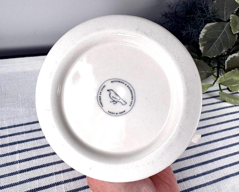 Op de afbeelding: Onderkant van een witte keramische mok met een cirkelvormig ontwerp. De onderkant van de mok heeft een klein, cirkelvormig embleem met een vogelillustratie en de tekst "MICROWAVE & DISHWASHER SAFE" en "MADE IN CHINA".
