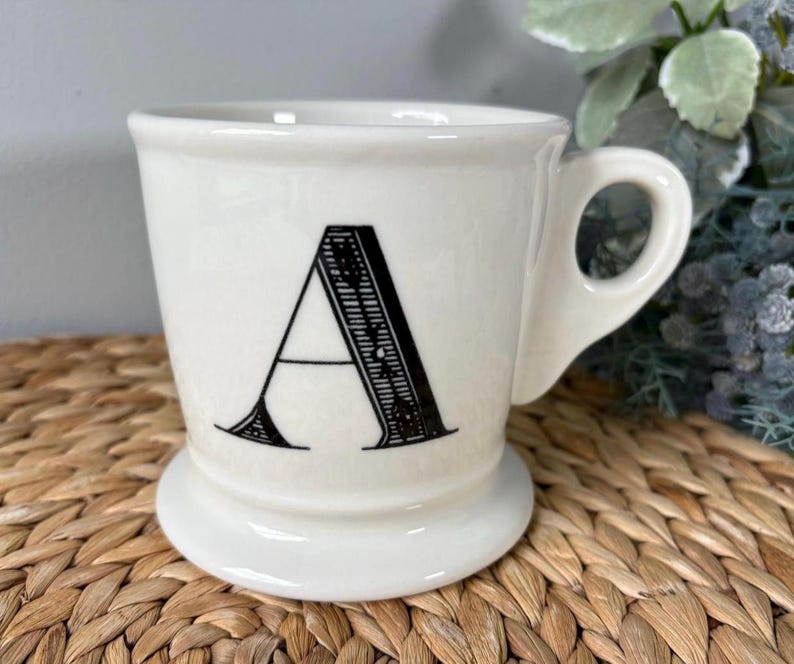 Pu&ograve; includere: Tazza in ceramica color crema con la lettera "A" nera sul davanti. La tazza ha una base arrotondata, un manico curvo e un bordo leggermente svasato. La lettera "A" &egrave; in un carattere decorativo in grassetto.