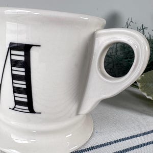 Op de afbeelding: Witte keramische mok met een grote zwarte letter "M" op de voorkant. De letter is gedeeltelijk gestreept. De mok heeft een uniek handvat en staat op een kleine voet. De mok staat op een gestreept oppervlak.