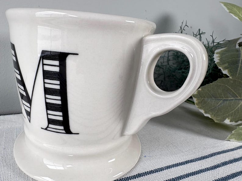 Op de afbeelding: Witte keramische mok met een grote zwarte letter "M" op de voorkant. De letter is gedeeltelijk gestreept. De mok heeft een uniek handvat en staat op een kleine voet. De mok staat op een gestreept oppervlak.