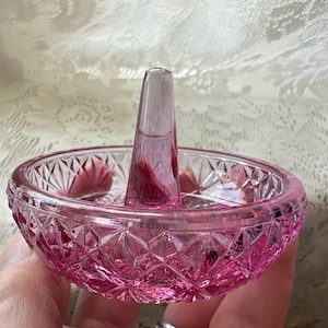 May include: A translucent pink glass ring holder featuring a circular dish base with a diamond-cut pattern and a central conical post. The glass has a vibrant magenta hue and a faceted exterior texture that catches the light across its surface.