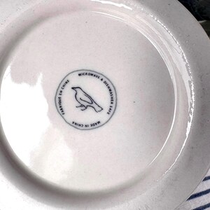Op de afbeelding: Witte keramische plaat met een licht gestructureerd oppervlak. De plaat heeft een cirkelvormig embleem met een vogelillustratie en de tekst "Microwave & Dishwasher Safe" en "Made in China".