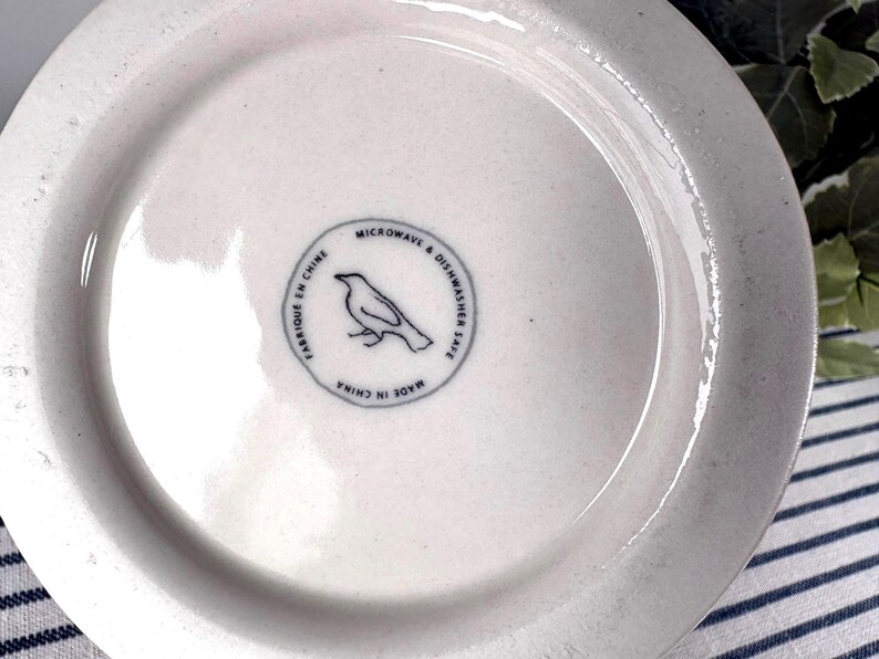 Op de afbeelding: Witte keramische plaat met een licht gestructureerd oppervlak. De plaat heeft een cirkelvormig embleem met een vogelillustratie en de tekst "Microwave & Dishwasher Safe" en "Made in China".