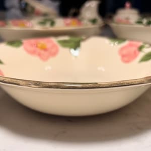 May include: Cream-colored ceramic bowl with a pink floral pattern and green leaves. The rim is trimmed with a gold-colored band. Other matching dishware is visible in the background.