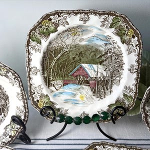 May include: A square ceramic plate featuring a central illustration of a red covered bridge over a stream in a snowy landscape. The border is decorated with brown and green embossed leaf and floral patterns on a white background.