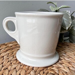 Pu&ograve; includere: Una tazza in ceramica bianca con un manico arrotondato e una base ampia e stabile. La tazza ha una finitura liscia e lucida e un design classico e semplice. La tazza viene probabilmente utilizzata per bere bevande.