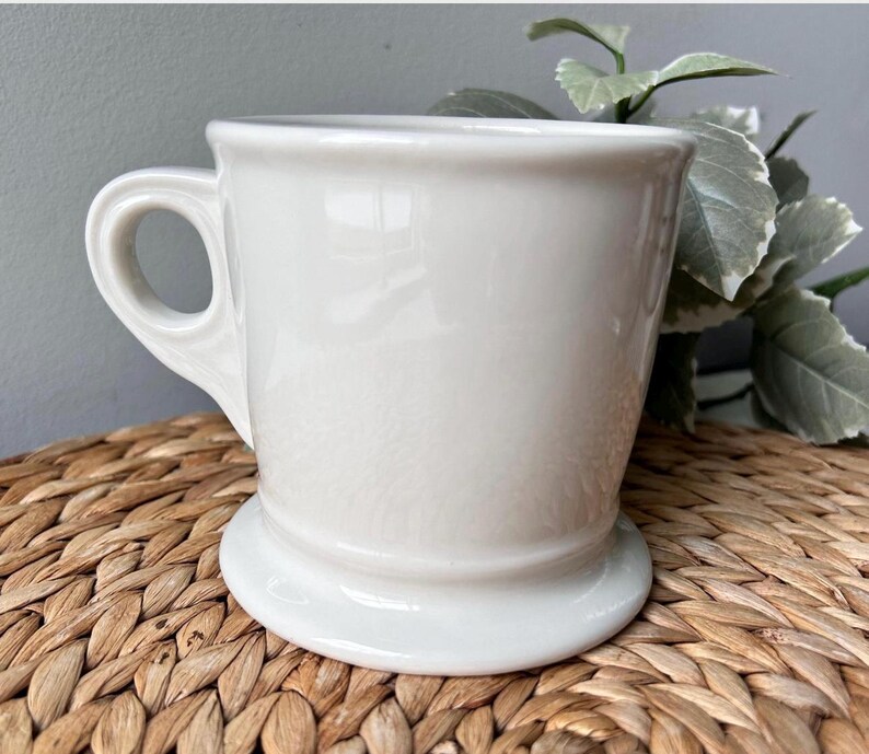 Pu&ograve; includere: Una tazza in ceramica bianca con un manico arrotondato e una base ampia e stabile. La tazza ha una finitura liscia e lucida e un design classico e semplice. La tazza viene probabilmente utilizzata per bere bevande.