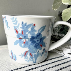May include: A white ceramic mug featuring a blue floral pattern with small red accents. The mug has a wide, slightly tapered body and a curved handle on the right side. The design consists of various blue flowers and leaves scattered across the surface.