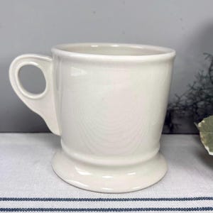 Op de afbeelding: Cr&egrave;mekleurige keramische mok met een afgeronde basis en een handvat. De mok heeft een eenvoudig, klassiek ontwerp, met een gladde, glanzende afwerking. De handgreep heeft een lusvorm. De mok staat op een wit en blauw gestreept oppervlak.