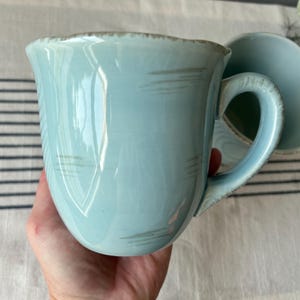 May include: A light blue ceramic mug with a curved handle and a slightly textured surface. The mug has a wide opening and a matching saucer is visible in the background. The mug has a capacity of approximately 12 ounces.