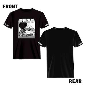 Op de afbeelding: Zwart T-shirt met een witte afbeelding van een auto die een berg op rijdt met de tekst "峠 伝説" op de voorkant. De mouwen hebben de tekst "TOUGE" en "NATION" erop gedrukt.