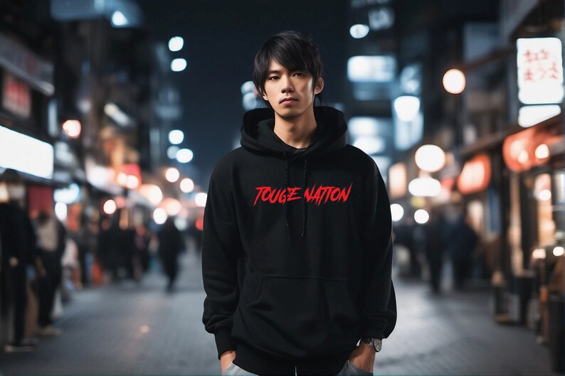 Puede incluir: Sudadera negra con el texto rojo "TOUGE NATION" en el pecho. La imagen est&aacute; tomada de noche en una calle de la ciudad con luces borrosas en el fondo. La sudadera tiene capucha y mangas largas.