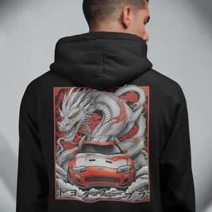 Puede incluir: Sudadera con capucha negra con un dise&ntilde;o de coche deportivo rojo y blanco y un drag&oacute;n blanco. El dise&ntilde;o incluye texto japon&eacute;s. El coche est&aacute; delante del drag&oacute;n, sobre un fondo rojo.