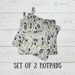 May include: Two square hot pads with hanging loops. The pads feature a white background with a repeating pattern of green and black leaves, and small pink dots. The text "SET OF 2 HOTPADS" is printed in black.