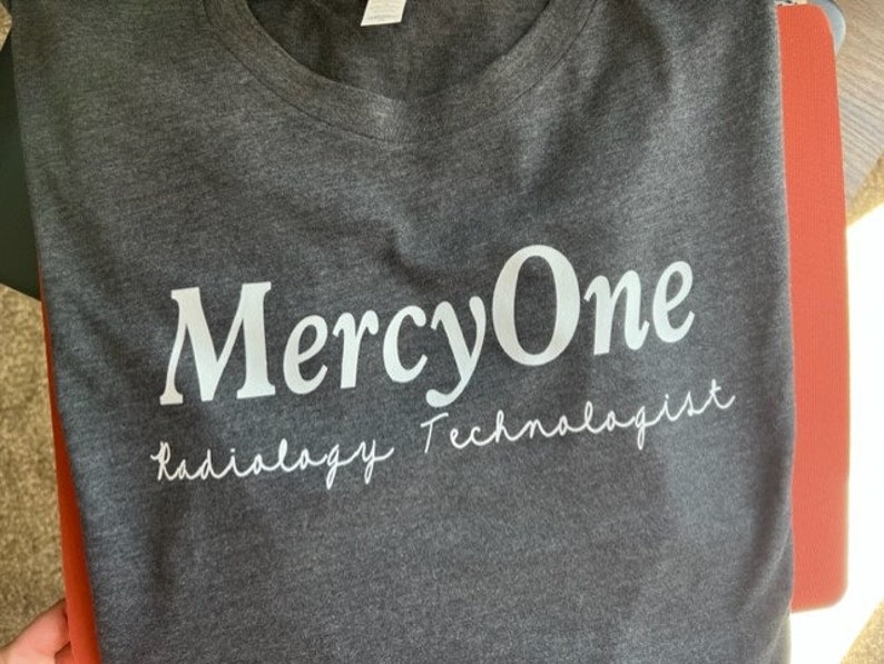 May include: A dark gray t-shirt with the text "MercyOne Radiology Technologist" printed in white.