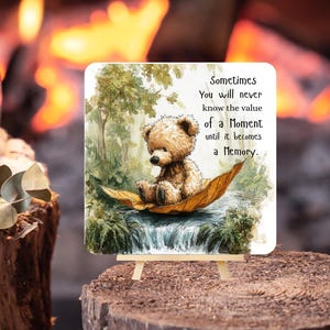 May include: A square decorative sign featuring a watercolor illustration of a brown teddy bear sitting on a large yellow leaf floating on a stream. The text reads: Sometimes You will never know the value of a Moment until it becomes a Memory.