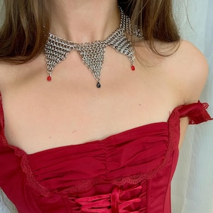 May include: A silver chainmail choker necklace with three pendant drops, two red and one black, worn with a red corset top. The corset has a lace-trimmed neckline and a ribbon-laced front.