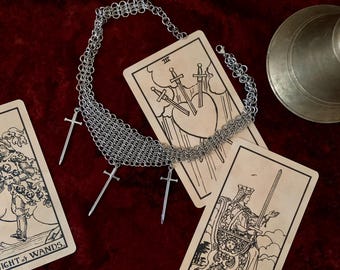 Könnte beinhalten: Eine silberne Kettenhemdkette mit vier silbernen Schwertpendants. Die Kette liegt auf drei Tarotkarten: Königin der Schwerter, Drei der Schwerter und Ritter der Stäbe.