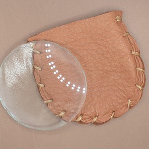 May include: A clear magnifying glass with a stitched leather case. The case is tan and has a rounded shape. The glass is partially covered by the case, which is stitched with a contrasting thread. The magnifying glass is a tool for reading.