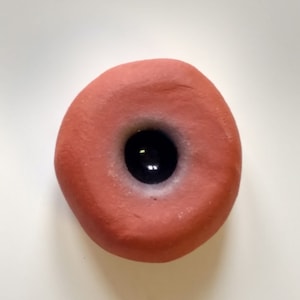 May include: A small, round, terracotta-colored ceramic object with a black, circular hole in the center.