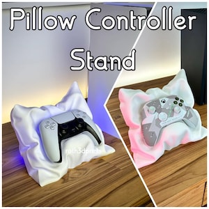 May include: A white 3D-printed pillow controller stand with a PlayStation 5 controller resting on it. The stand is on a wooden surface. The text "Pillow Controller Stand" is at the top of the image.