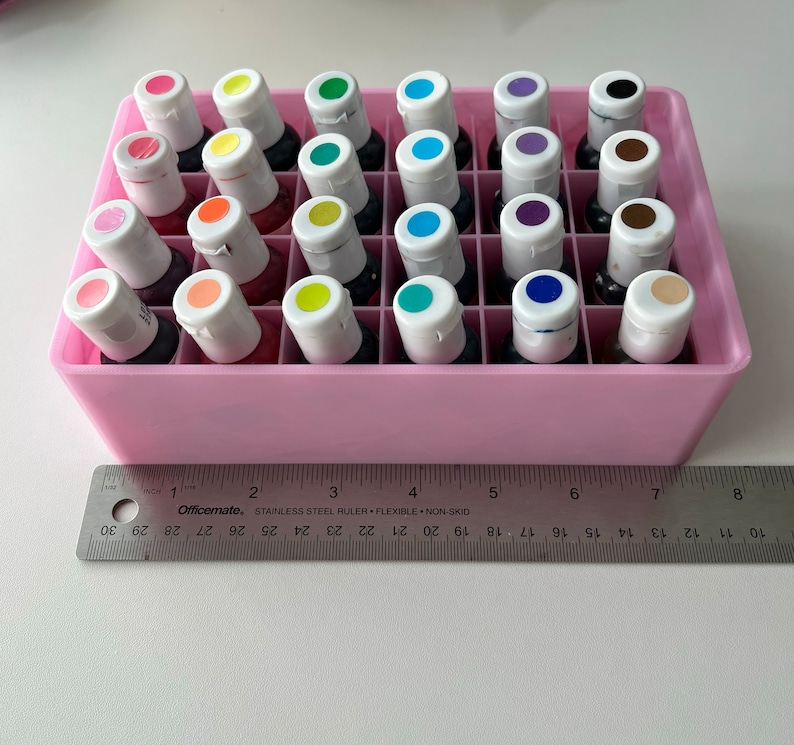 May include: A pink plastic storage case with 24 small bottles of colorful liquid. The bottles have white caps and are arranged in a grid pattern.