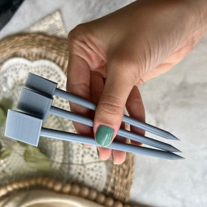 May include: Three gray plant markers with rectangular labels and pointed tips are held in a hand. The markers are made of a light gray material and are designed for use in a garden or with potted plants.