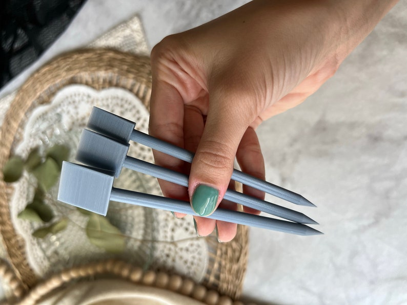 May include: Three gray plant markers with rectangular labels and pointed tips are held in a hand. The markers are made of a light gray material and are designed for use in a garden or with potted plants.