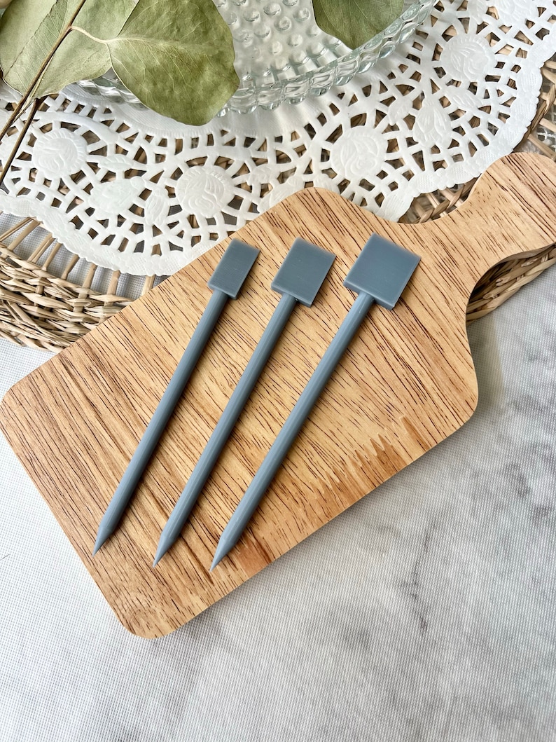May include: Three gray, pointed skewers with square tops rest on a wooden cutting board. The skewers are arranged diagonally, with the points facing down. The cutting board has a natural wood grain pattern. The background includes a white doily and dried greenery.