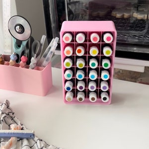 May include: A pink plastic storage organizer with 24 clear bottles containing colorful liquids. The bottles are arranged in a grid pattern and are labeled with numbers.