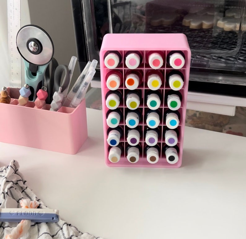 May include: A pink plastic storage organizer with 24 clear bottles containing colorful liquids. The bottles are arranged in a grid pattern and are labeled with numbers.